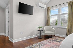 A room with a flat-screen TV mounted on the wall, an air conditioning unit, a window with blinds, a chair, a small table with a plant, and a patterned rug. - The Roosevelt Suite | Germantown Inn | Breakfast (Nashville)