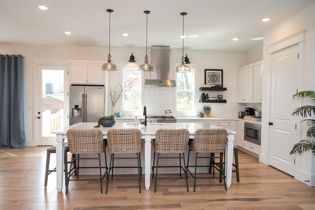 Una cocina moderna con una isla central, cuatro taburetes de bar, iluminación colgante y un refrigerador de acero inoxidable.