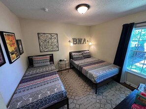 A room with two beds, a small round table, a wall-mounted light, and a window with curtains. - 3BR Cottage - 3 Miles to Downtown (Nashville)