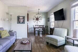 A living room with a sofa, coffee table, patterned ottoman, dining area with chairs, a wall-mounted TV, and a large compass decoration. - 3BR Cottage - 3 Miles to Downtown (Nashville)
