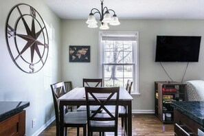 A dining area with a wooden table and chairs, a wall-mounted compass, a window with blinds, and a television on the wall. - 3BR Cottage - 3 Miles to Downtown (Nashville)
