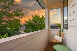 Un balcon avec une chaise en osier et une petite table, d’où l’on peut admirer un coucher de soleil avec des arbres et des bâtiments en arrière-plan.