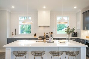 A modern kitchen with white cabinetry, a large island with bar stools, pendant lighting, and a view of greenery outside. - Chic 3BR + Free 2-Car Garage | Near Downtown (Nashville)