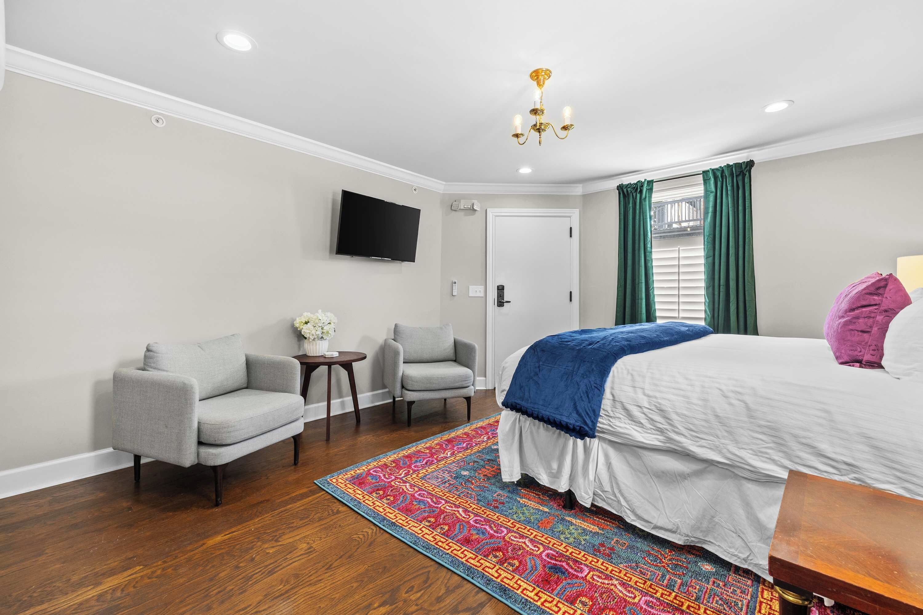 A hotel room with a bed, two armchairs, a small table with a vase, a television on the wall, and a colorful rug.