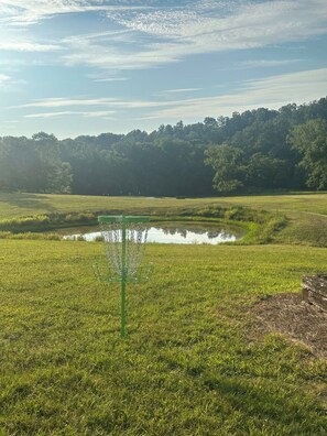 Un campo de disc golf con un cesto de disc golf verde en primer plano, un estanque en el centro y una colina boscosa al fondo.