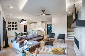 Ein modernes Wohnzimmer mit einer Couch, einem Couchtisch und Sesseln, eine Küche im Hintergrund und eine Treppe, die in eine obere Ebene führt.
