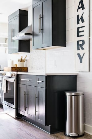 A modern kitchen with dark cabinetry, a stainless steel trash can, and a framed word "BAKERY" on the wall. - Elegant Farmhouse 15 Min to Nashville (Nashville)