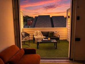 Een balkon met een bank, een stoel en een tafel, met uitzicht op een woonwijk tijdens zonsondergang.