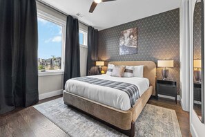 A bedroom with a large bed, two bedside lamps, a window with curtains, a city view, and a patterned wall. - Bourbon & Ballads: Karaoke Stage, Sauna + Rooftop (Nashville)