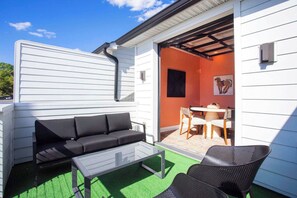 Un patio con un sofá negro, una mesa de centro de vidrio y dos sillas de mimbre.
