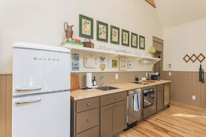 A compact kitchen with a Galanz refrigerator, wooden cabinets, a sink, and a dishwasher. There are framed botanical prints on the wall and a shelf with dishes and a vase. - The Roost at Buckhorn Hollow (McEwen)