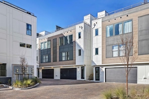 A modern residential building with multiple garage doors, a brick driveway, and a small tree in the foreground. - City Views | Rooftop Deck | Fire Pit + Grill (Nashville)