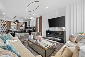 A modern living room with a glass coffee table, sofas, a TV, and a kitchen area visible through an open door. - City Views | Rooftop Deck | Fire Pit + Grill (Nashville)