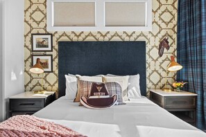 A neatly made bed with a decorative pillow, a nightstand with a lamp, and a patterned wall. - City Views | Rooftop Deck | Fire Pit + Grill (Nashville)