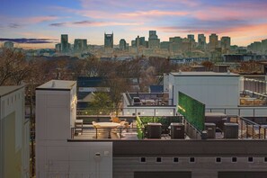 A rooftop terrace with outdoor seating, air conditioning units, and a view of a city skyline at sunset. - City Views | Rooftop Deck | Fire Pit + Grill (Nashville)