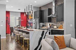 A modern kitchen with a central island, bar stools, and a grey sofa with an orange pillow. - Elegant Nash Karaoke Haven w/ Rooftop Fire Table (Nashville)