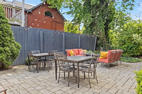 An outdoor patio with a brick floor, a wooden table with chairs, a sofa with cushions, and a wooden fence. - The Madison Suite | Germantown Inn | Breakfast (Nashville)