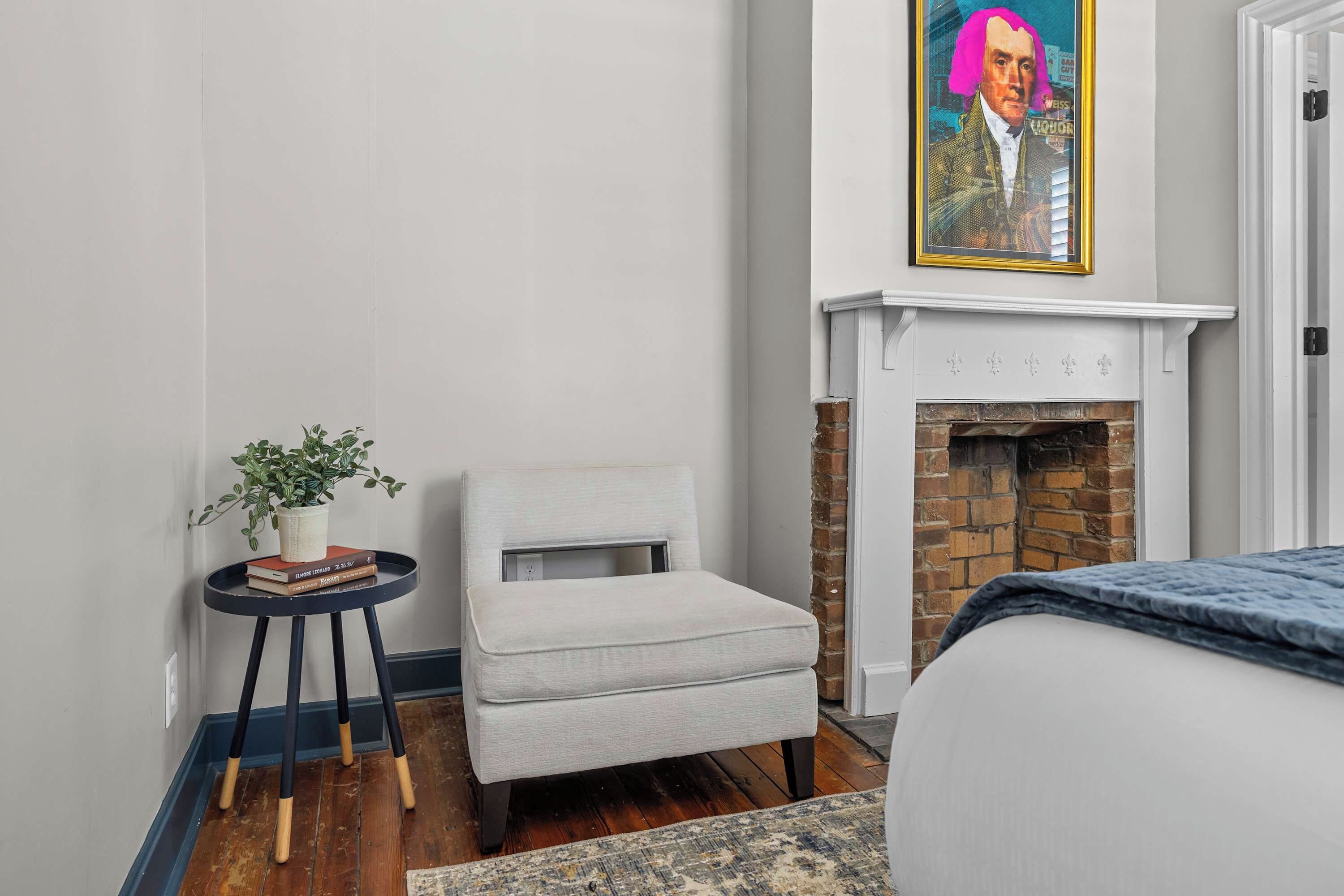 A bedroom with a bed, a small chair, a side table with a plant, a fireplace, and a framed picture on the wall.