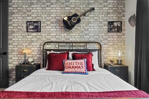 Una cama con sábana blanca, almohadas rojas y una almohada con el texto “¿SOY EL DRAMA?”. Hay una guitarra en la pared y dos cuadros enmarcados.