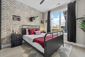 Quarto com parede de tijolos, cama com almofadas vermelhas e azuis, um violão e uma janela com cortinas.