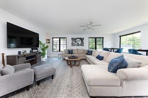 A modern living room with a large flat-screen TV, a sectional sofa, a coffee table, and a ceiling fan. - New Hot Tub, Spacious, Near Downtown (Nashville)