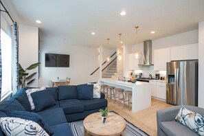 A modern living room with a sectional sofa, a round coffee table, and a grey sofa. A kitchen with a stainless steel refrigerator and bar stools is visible in the background. - 2 Side by Side Homes with Rooftops -Sleeps 24 (Nashville)