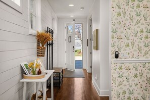 A hallway with a white door, a small table with a vase and a book, and a wall with a floral pattern. - Spotless Gem | Hot Tub | 15 Min to Broadway (Nashville)