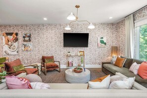 Ein modernes Wohnzimmer mit strukturierter Tapete, einem Flachbildfernseher und verschiedenen Sitzgelegenheiten, darunter eine Couch, Sessel und ein runder Couchtisch.