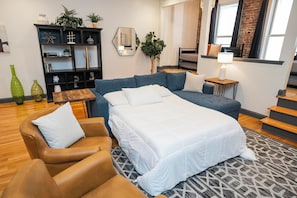 A modern bedroom with a large bed, a sofa, a chair, and a small table. There is a bookshelf with decorative items and a plant. - On Broadway Above Boot Barn (Nashville)