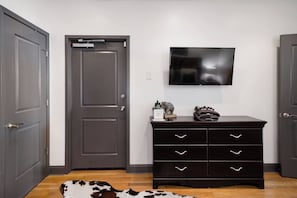 A room with a dark wooden dresser, a flat-screen TV mounted on the wall, and a pair of closed doors. - On Broadway Above Boot Barn (Nashville)