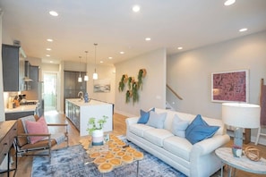 A modern living room with a sofa, coffee table, and a kitchen area in the background. - 2 Homes-Sleeps 24-Roof Decks-Grills Near Broadway (Nashville)