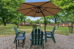Eine Terrasse im Freien mit Tisch und Stühlen unter einem großen Sonnenschirm, umgeben von Bäumen und einem Pflasterweg.