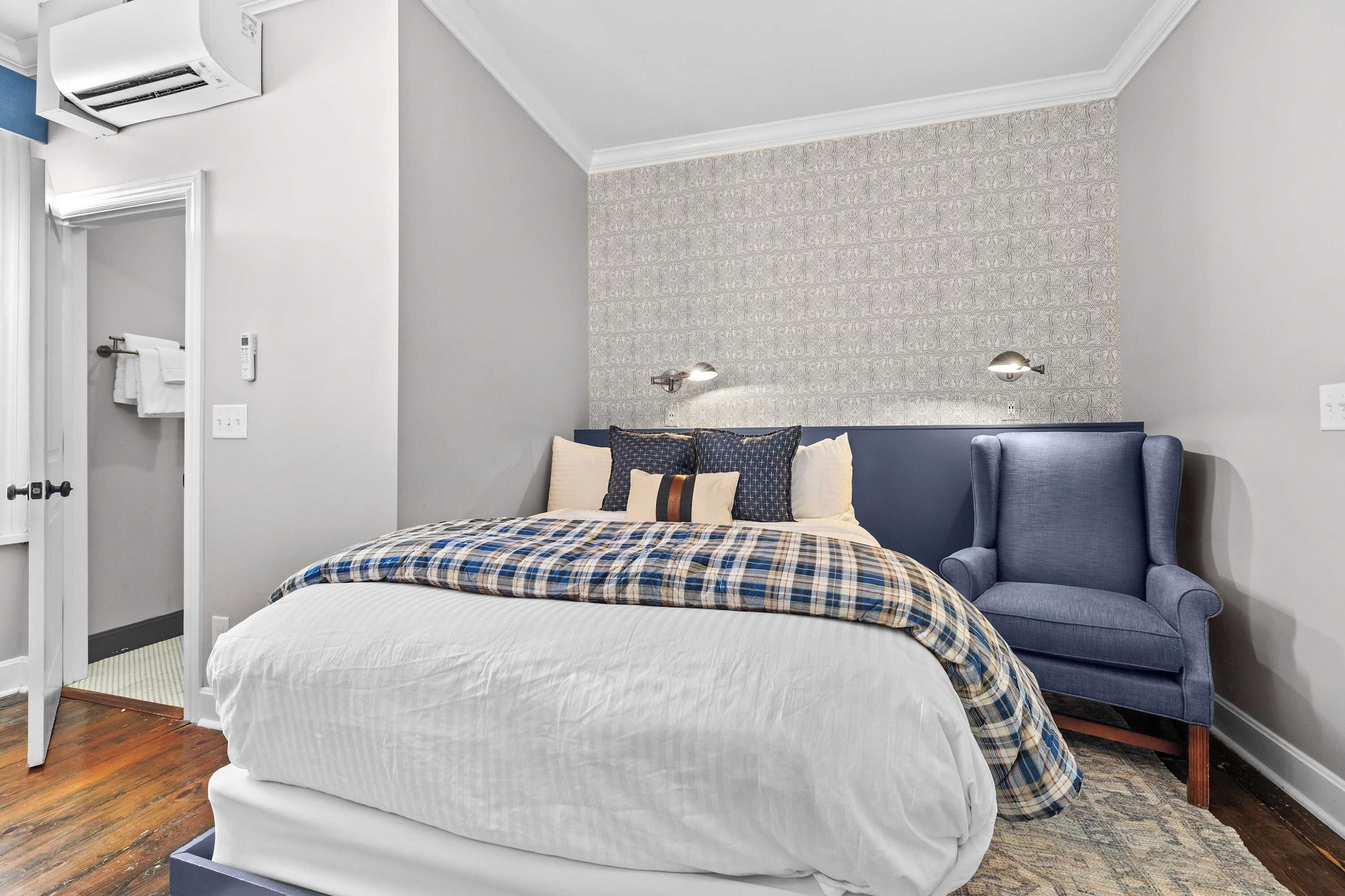 A bedroom with a bed, a chair, a wall-mounted lamp, and a patterned wallpaper.