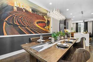A dining area with a long wooden table set for a meal, a mural of an auditorium on the wall, and a modern chandelier. - Karaoke, Rooftop, Fire Pit -The Ryman (Nashville)