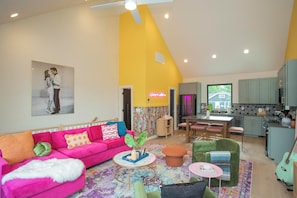 A modern living room with a vibrant yellow accent wall, a pink sofa, a green armchair, and a kitchen area with a dining set. - Bohemian Chic Hot Tub Sleeps 12 (Nashville)