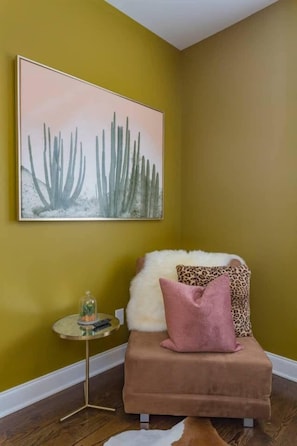 Ein Zimmer mit gelber Wand, einer gerahmten Kakteenkunst, einem kleinen runden Tisch mit einer Glaskuppel und einem braunen Sofa mit gemusterten Kissen.