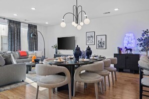 A modern living room with a dining area, featuring a large flat-screen TV, a chandelier, and a neon sign. - Nash Skyline Views Rooftop with Fire Pit  Grill (Nashville)