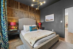 Quarto com cama grande, painel de parede em madeira, duas mesas de cabeceira com luminárias, um quadro na parede e uma porta que dá para outro cômodo.