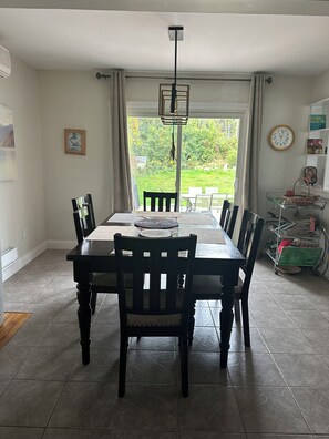 A dining area with a table and chairs, a hanging light fixture, a window with curtains, and a view of greenery outside. - Dual Purpose Comfort: 4BR Park & Beach House + Office & High Speed WiFi (Kawartha Lakes)
