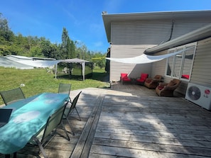 Une terrasse extérieure couverte avec une table et des chaises, un espace salon couvert avec des coussins rouges et un appareil de climatisation.