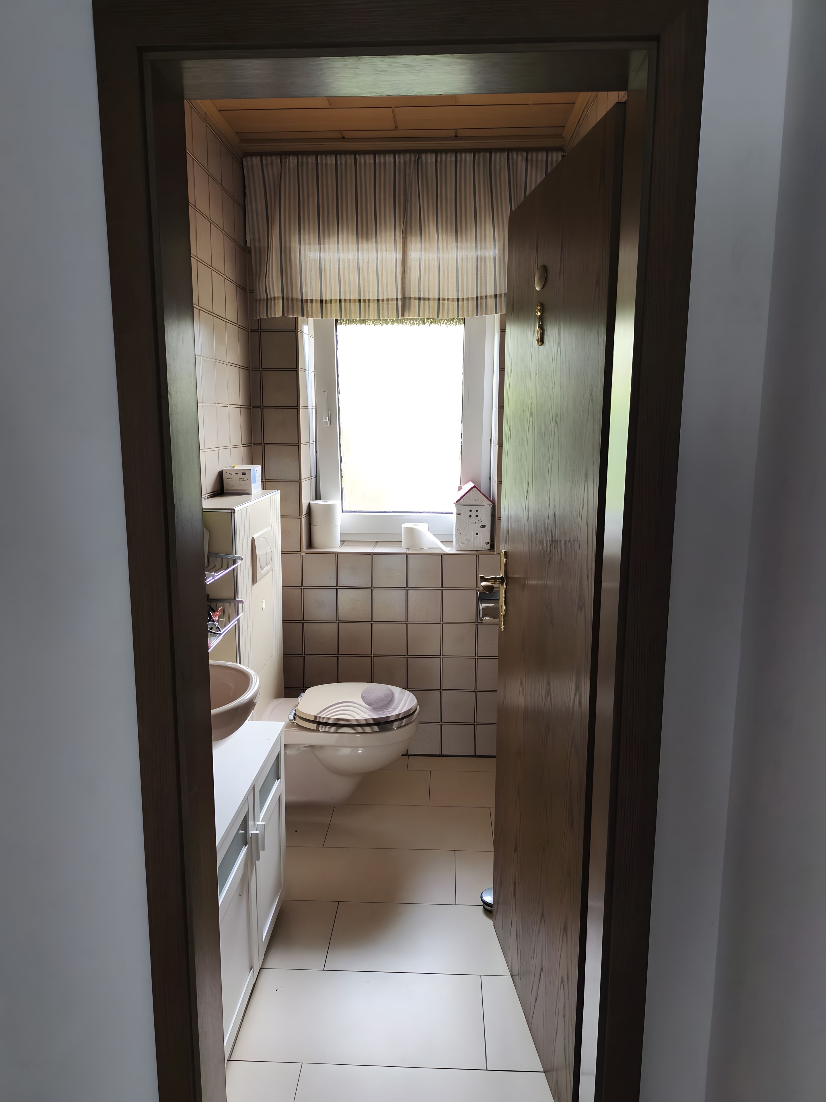 Een smalle gang die leidt naar een badkamer met toilet, wastafel en raam.