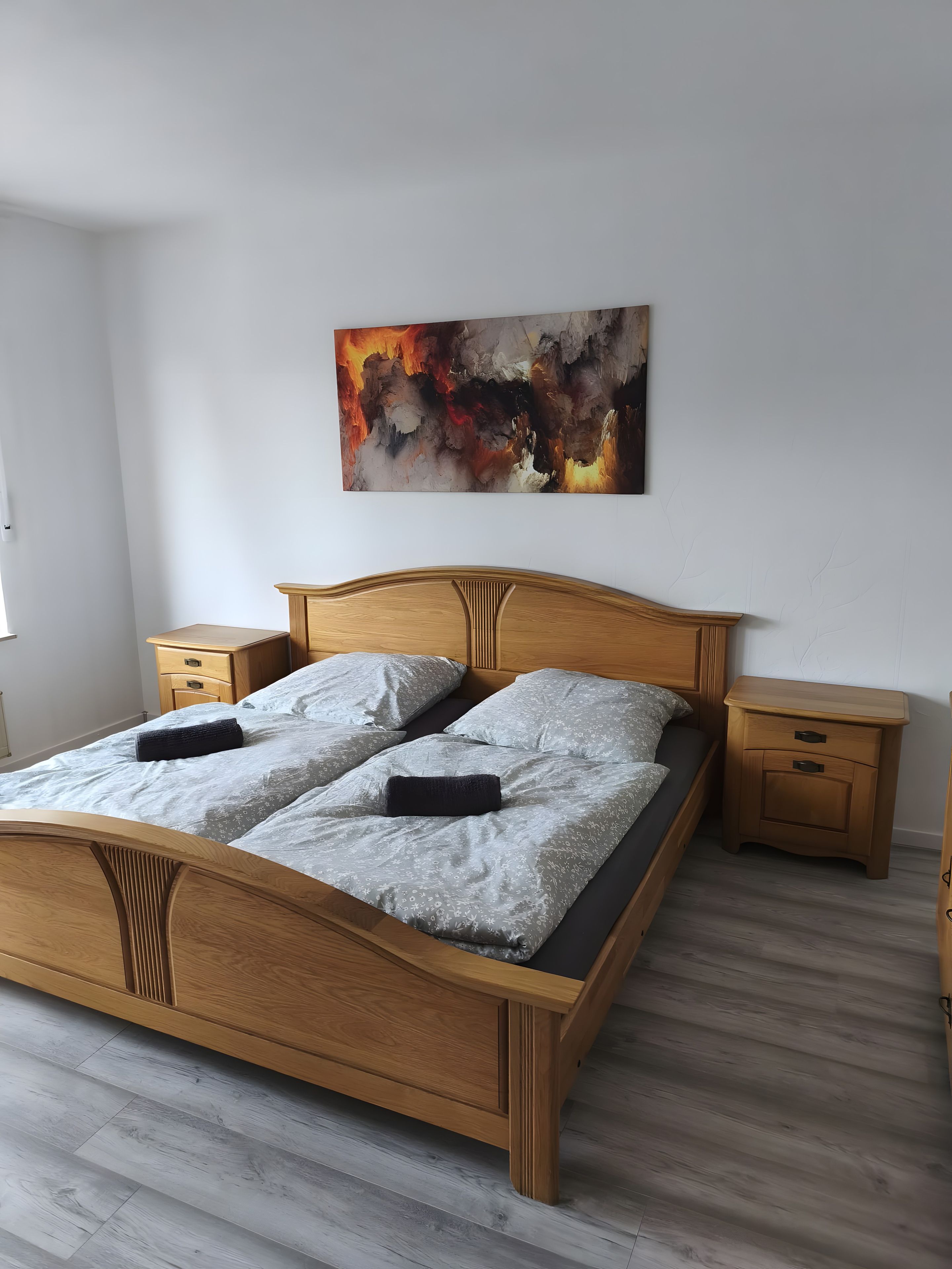 A bedroom with a wooden bed, grey patterned bedding, a wooden nightstand with drawers, and a wall-mounted abstract painting.