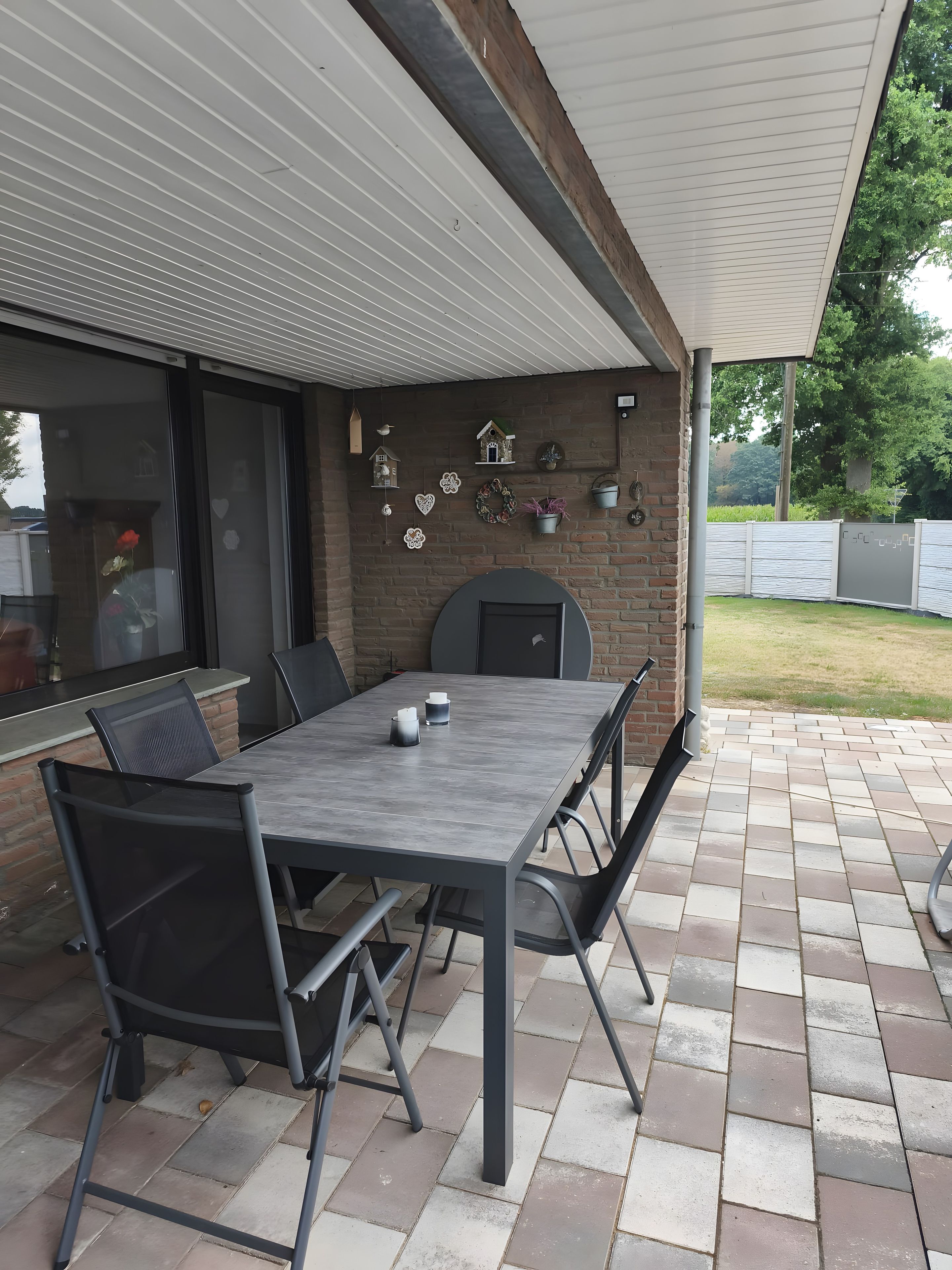 Un patio al aire libre con una mesa y sillas, un muro de ladrillo con adornos y una zona cubierta.
