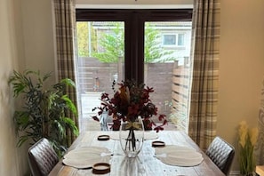 A dining table with a vase of flowers, surrounded by chairs, with a view of a building and greenery outside. - House Sleeps 4 With Enclosed Garden (Oban)