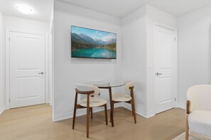 Ein Zimmer mit weißer Tür, weißer Wand, einem Wand-Fernseher, der eine Berglandschaft zeigt, einem runden Glastisch mit zwei Stühlen und Holzboden.