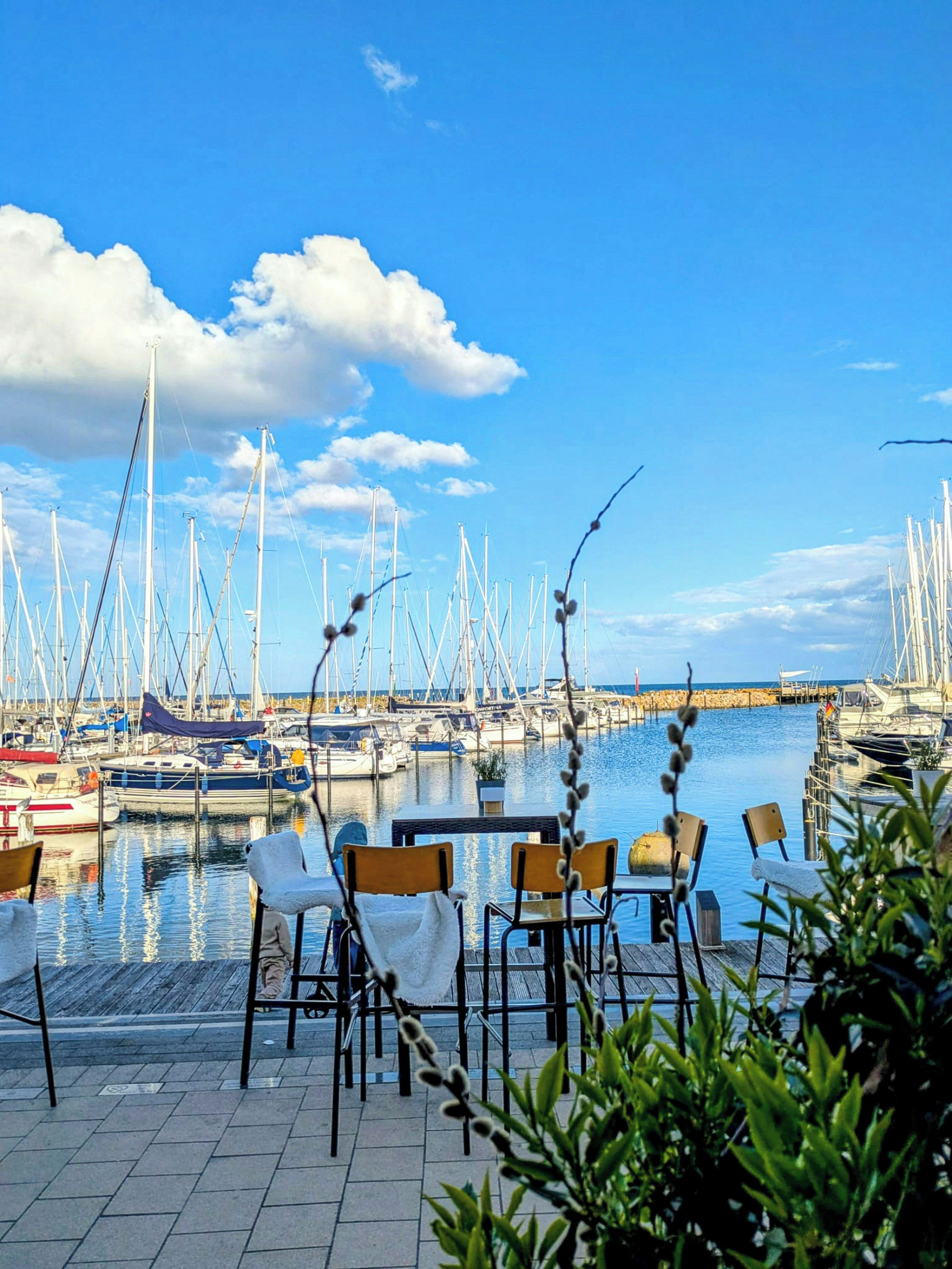 Un port de plaisance avec des bateaux amarrés, un ciel dégagé et des espaces de détente en plein air avec des chaises et des tables.