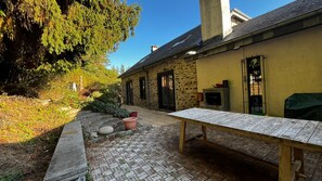 A courtyard with a stone floor, a wooden table, and a stone building with windows and a chimney. - Cozy vacation home with a private forest, surrounded by nature and close to Houffalize. (Houffalize)