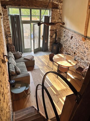A cozy interior with a stone wall, a wooden staircase, a sofa, a dining table, and a view through a glass door.