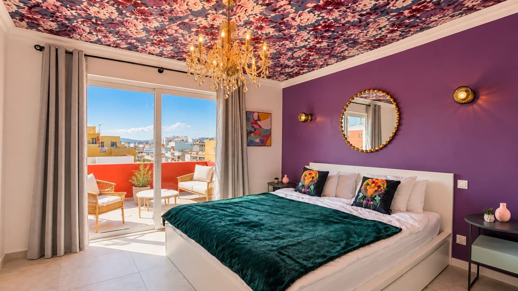 A bedroom with a large bed, a chandelier, a balcony with a view, and a purple wall.