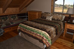 Un dormitorio con techo estilo loft, una cama con ropa de cama estampada, una mesita de noche, una lámpara y vistas al exterior.